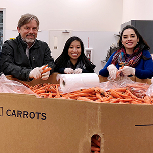 3 employees at the food bank volunteering, standing in front of box of carrots